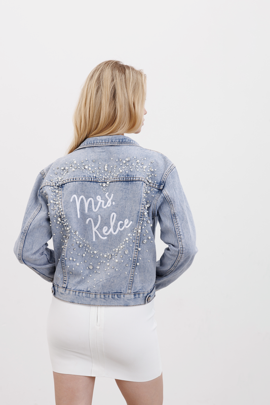 A woman facing away from the camera wears a blue denim jacket adorned with crystals, faux pearls, and rhinestones forming a heart shape on the back, along with the personalized text 'Mrs. Keia'.