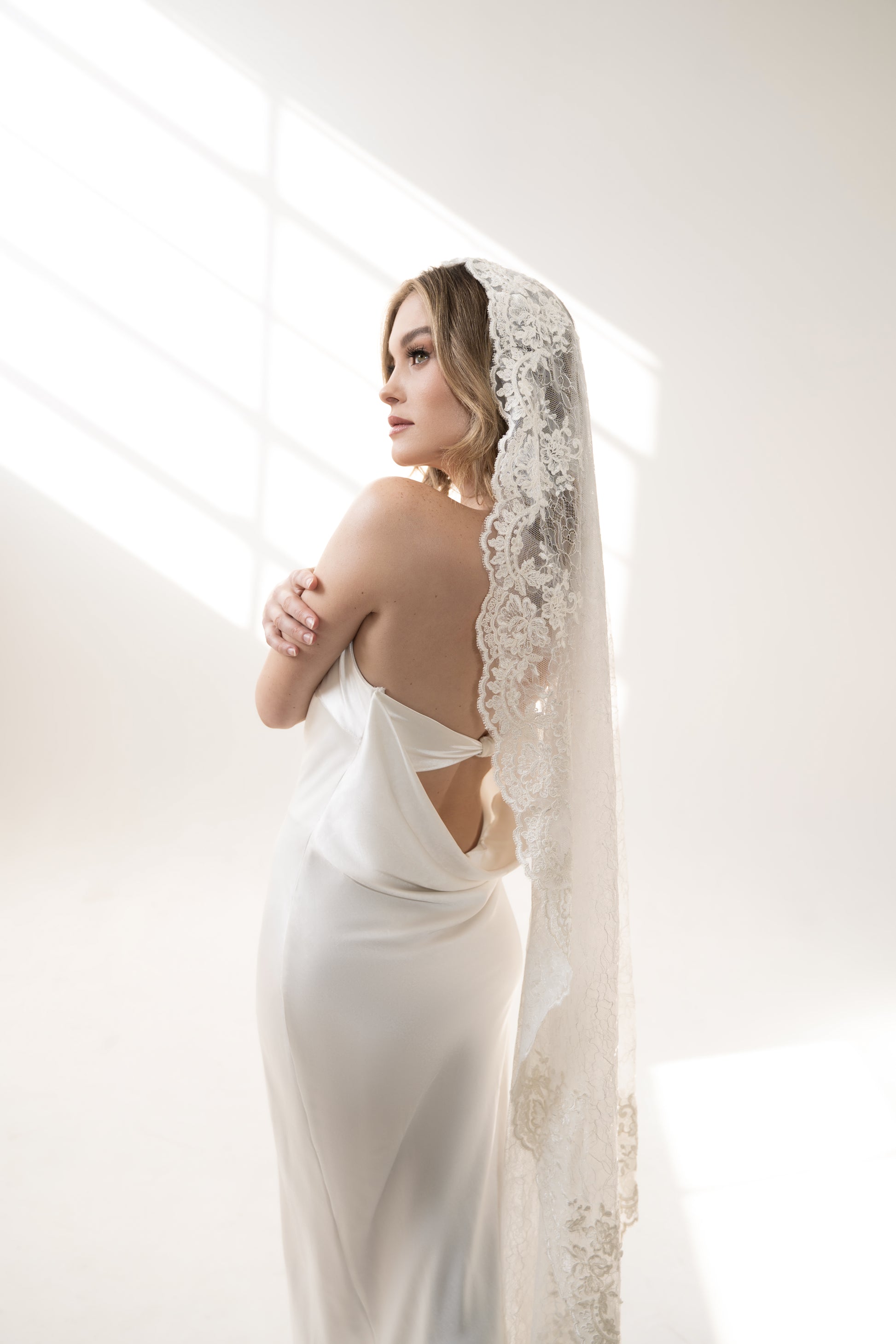 A model wearing a white, mantilla-style bridal veil with Chantilly lace detailing, in a waltz length that extends to the floor.